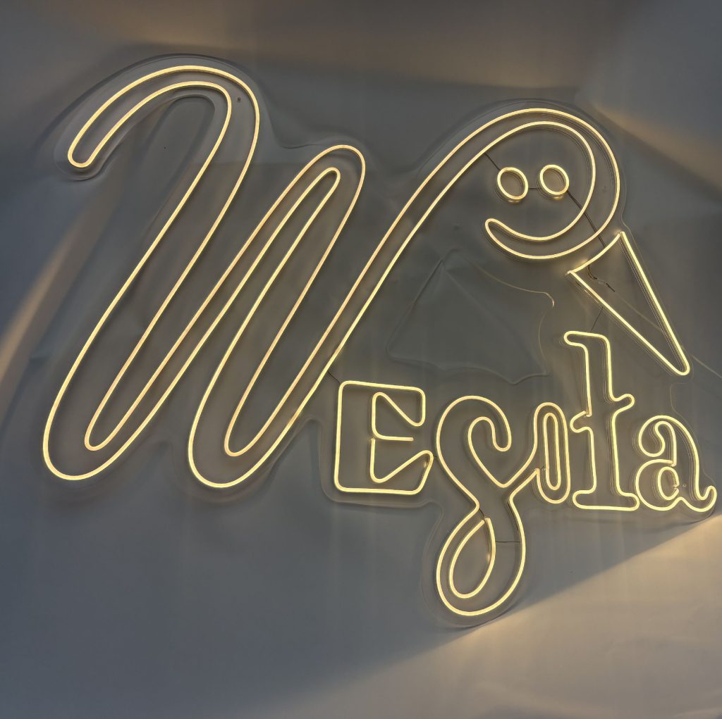Świecący ciepłym światłem neon LED z napisem Wesoła wkomponowanym w bociana, wykonany na podkładzie z bezbarwnej pleksi w Zalasewo.