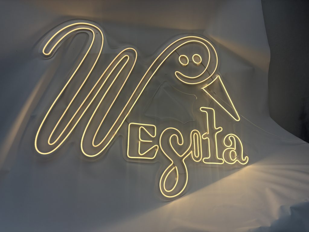Świecący ciepłym światłem neon LED z napisem Wesoła wkomponowanym w bociana, wykonany na podkładzie z bezbarwnej pleksi w [Zalasewo].