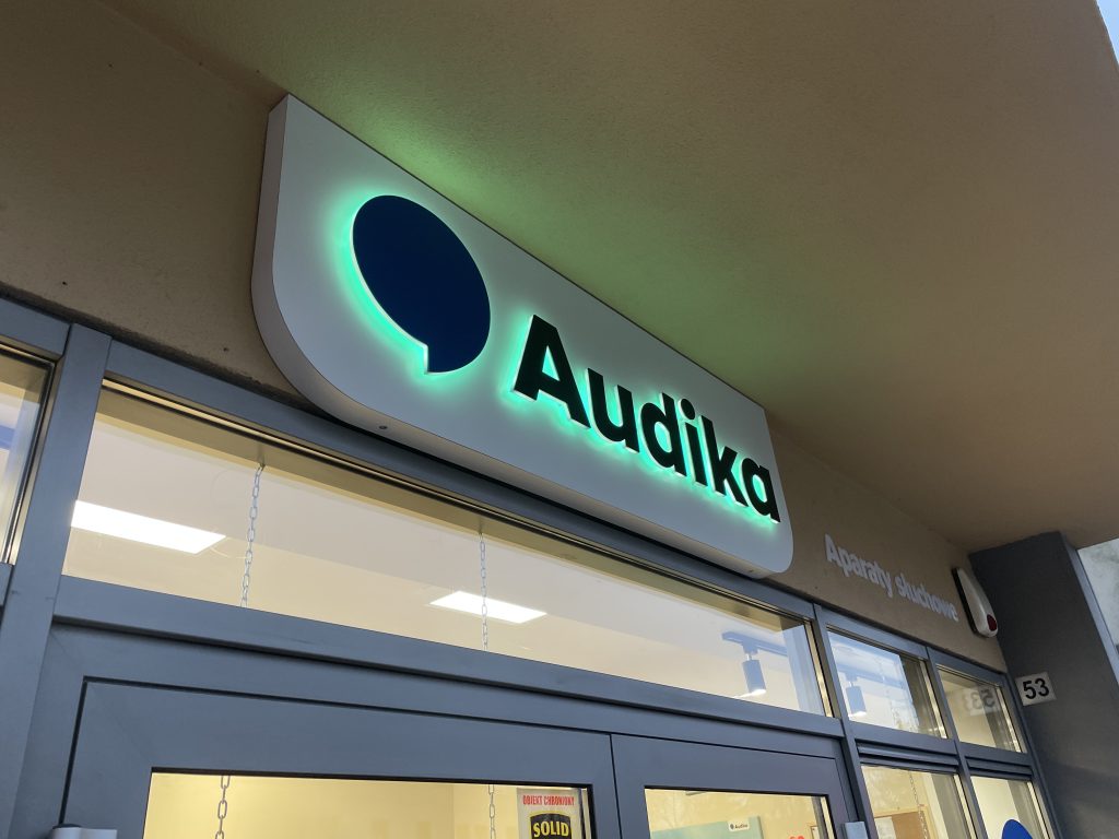 Szyld reklamowy z literami 3D podświetlanymi od tyłu (efekt halo) na zielono, zamontowany na białym panelu maskującym nad wejściem do salonu w [Swarzędz].